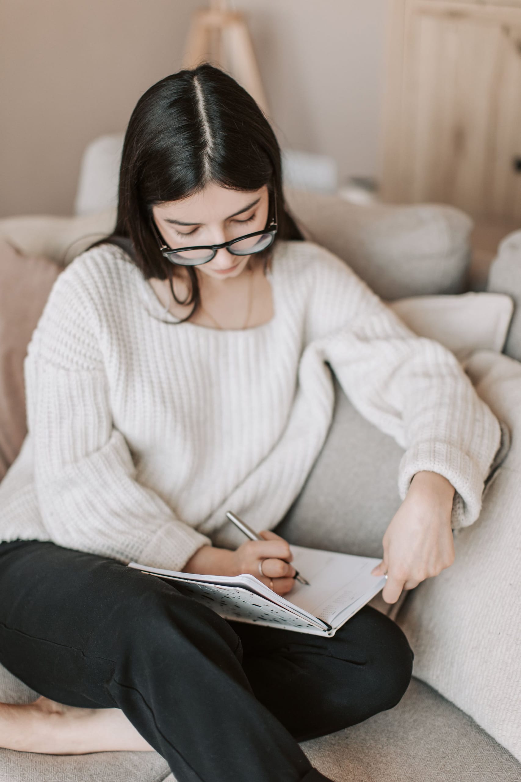 A woman in a white sweater writing in a notebook on the sofa