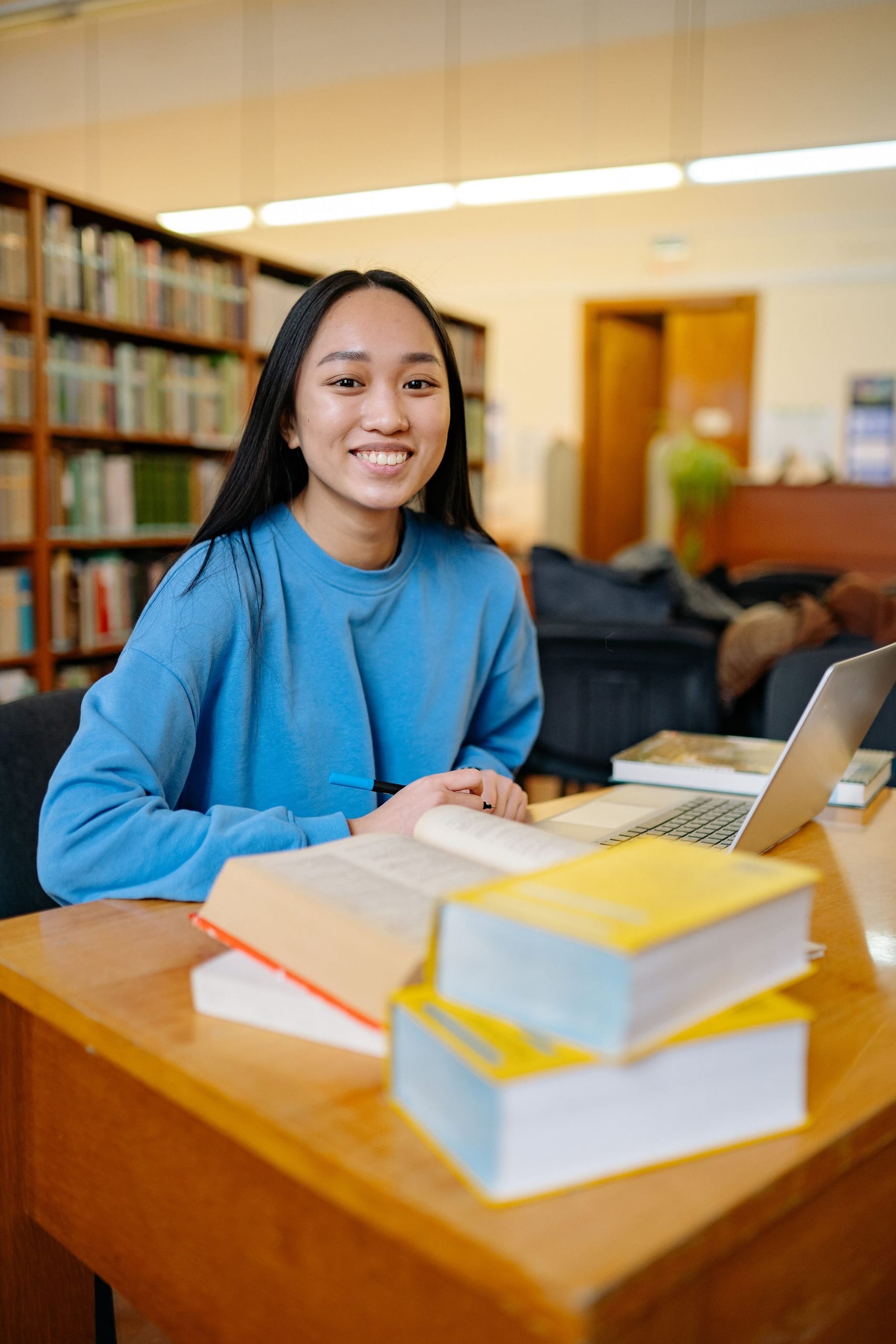 A smiling student studying at a library table with books and a laptop